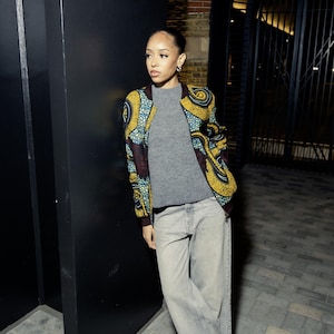 May include: A woman wearing a grey sweater, blue jeans, and a colourful patterned bomber jacket. She is standing next to a black wall.