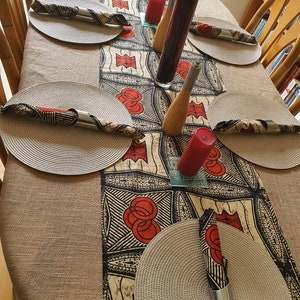 May include: A table setting with a black and white patterned table runner with red accents. The table runner is placed on a beige tablecloth. There are four place settings with woven placemats and napkins. There are also two candlesticks with red candles and a glass vase with a black and white patterned design.