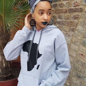 Puede incluir: Una sudadera gris con un contorno negro del continente africano en la parte delantera. La sudadera tiene una capucha con cordón y un bolsillo canguro. La persona que lleva la sudadera también lleva una cinta para la cabeza con estampado blanco y negro y grandes pendientes redondos que dicen "Faith Love Hope".