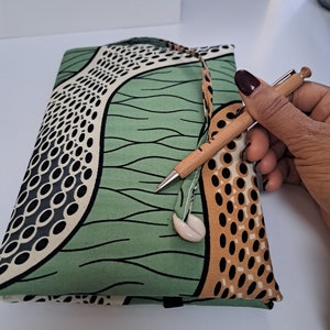 May include: A closed, rectangular journal with a green, tan, and black patterned fabric cover. A wooden pen rests on the journal. A seashell charm is attached to the journal's closure. The journal has a handle.