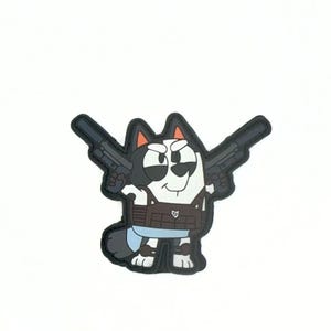 Könnte beinhalten: Ein Cartoon-Patch mit einem Hund mit schwarz-weißen Markierungen, der eine braune Weste trägt und zwei schwarze Waffen hält. Der Hund hat einen ernsten Gesichtsausdruck. Der Patch hat einen schwarzen Rand auf weißem Hintergrund.