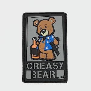 Op de afbeelding: Een rechthoekige patch met een cartoonbeer in een blauw Hawaiiaans shirt, met een fles en een geweer. De beer is bruin met een grijze achtergrond. De tekst "CREASY BEAR" staat onderaan in witte letters.