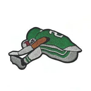 May include: Embroidered patch featuring a football player in a green uniform with white stripes and the number 2. The patch has a gray border and depicts the player in a kneeling position, with a brown arm and a red football.