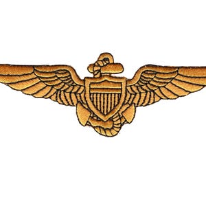 Puede incluir: Parche bordado de color dorado que representa un escudo con líneas verticales, un ancla y alas. Las alas están extendidas y el parche tiene un aspecto detallado y texturizado. El diseño es simétrico y centrado.