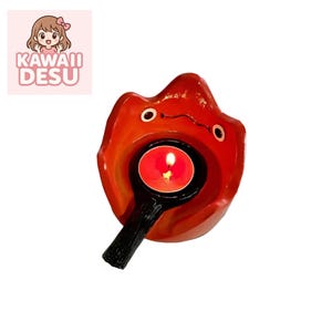 Könnte beinhalten: Ein roter Kerzenhalter aus Keramik in Form einer Flamme mit einem schwarzen Griff. Der Kerzenhalter hat zwei kleine, runde Augen und eine brennende rote Kerze in der Mitte. Der Text "KAWAII DESU" befindet sich in der oberen linken Ecke.