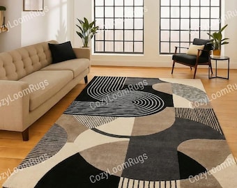 May include: A large, rectangular area rug with a geometric design. The rug features a mix of black, white, and gray shapes, including curved lines, circles, and rectangular blocks. The rug is placed on a hardwood floor in a well-lit room.