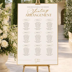 May include: A white wedding seating arrangement sign with gold lettering and a gold frame, displayed on a gold metal easel. The sign features the text 'YOUR SPECIAL PLACE AT OUR WEDDING', 'Seating ARRANGEMENT', and 'Olivia & John', with nine tables listed.