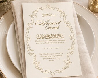 May include: A cream-colored rectangular wedding card featuring gold foil calligraphy and an ornate border. The text reads Ahmed & Sarah, followed by Arabic script and the English blessing: May Allah bless you both, shower His blessings upon you, and unite you both in goodness. The card is placed on a folded beige linen napkin.