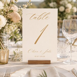 May include: A rectangular white table number card featuring the gold text table 1, the names Olivia & John, and the date 9/07/2030. The card is held upright by a small, rectangular wooden base, displaying elegant gold calligraphy and sans-serif font.