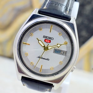 May include: Silver Seiko 5 automatic watch with a white face, gold hands, and a black leather band. The watch face displays the day and date, with the day reading "SAT 31". The Seiko logo is red.