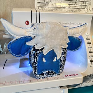May include: A handmade fabric cow head with blue and white accents. The cow head features large blue ears, a patterned blue and white face, and white horns. The cow head is placed on a white sewing machine.