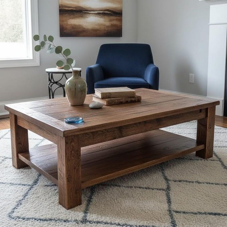 May include: A rectangular, wooden coffee table with a lower shelf. The table is a warm brown color and has a rustic, textured appearance. A vase, books, and a small decorative item sit on top. The table is in a living room setting.
