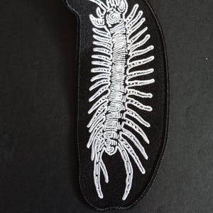 Peut inclure: Un écusson brodé noir avec un motif de mille-pattes blanc. Le mille-pattes a un corps segmenté avec de nombreuses pattes et antennes. L'écusson est de forme ovale et est probablement destiné aux vêtements ou aux accessoires.