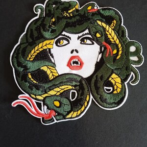 Könnte beinhalten: Ein gestickter Aufnäher mit einer stilisierten Darstellung der Medusa mit weißem Gesicht, roten Lippen und gelben Augen. Ihr Haar besteht aus dunkelgrünen und gelben Schlangen mit roten Zungen, die mit weißem Garn auf schwarzem Grund umrandet sind.