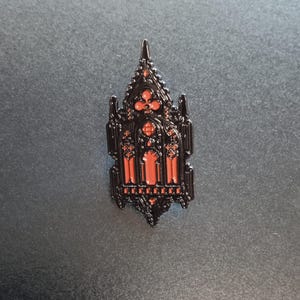 Peut inclure: Une broche en émail noir en forme d'arche de fenêtre gothique pointue. Le design présente des détails complexes en filigrane de métal noir qui encadrent des sections en creux remplies d'une couleur émail orange-rouge vive et unie.