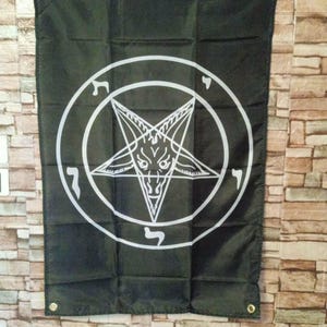 Op de afbeelding: Zwarte vlag met een witte pentagram en een geitenkop in een cirkel. De vlag heeft ogen in elke hoek om op te hangen. Het ontwerp is gecentreerd op de zwarte stof.