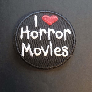 Peut inclure: Un écusson rond noir avec le texte brodé blanc "I heart Horror Movies". Un cœur rouge est brodé au-dessus du mot "Horror". L'écusson a une bordure noire.