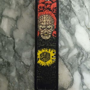 May include: Black rectangular keychain featuring a horror movie character with a red background of skulls. The design includes a yellow cube and a silver key ring.