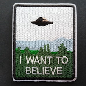 Peut inclure: Un écusson brodé rectangulaire représentant un OVNI noir planant au-dessus d'une forêt verte et de montagnes bleu clair sur un ciel blanc. Le texte I WANT TO BELIEVE est brodé en lettres capitales blanches sur la partie verte inférieure.