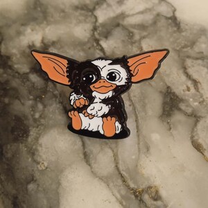 Op de afbeelding: Een kleurrijke emaille pin met een cartoon Gremlin-personage. De pin heeft een zwart-wit gezicht, grote oranje oren en zit met gekruiste armen en benen. De pin is een verzamelobject.