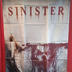 Op de afbeelding: Een rechthoekige stoffen wandbanner met de filmtitel SINISTER in rode, druipende letters. Daaronder staat een persoon met lang haar voor een muur bedekt met donkerrode verfspatten die de vorm van een schedel aannemen.