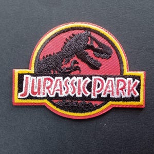 Peut inclure: Un écusson brodé circulaire présentant la silhouette noire d'un squelette de dinosaure sur fond rouge. Une bande noire horizontale au centre affiche le texte blanc JURASSIC PARK avec un contour bordé de jaune et de rouge.
