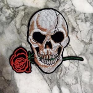 Peut inclure: Écusson brodé représentant un crâne avec un front fissuré et une rose rouge dans la bouche. Le crâne est blanc cassé avec des orbites noires et un contour noir. La rose est rouge avec des feuilles vertes et un contour noir.
