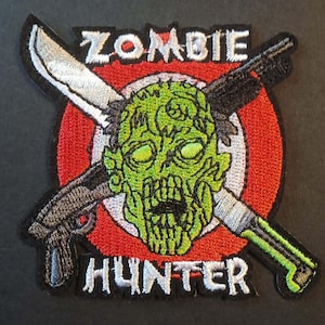 Peut inclure: Écusson brodé avec une tête de zombie verte, une machette et un fusil croisés derrière. L'écusson est posé sur une cible rouge et blanche, avec les mots "ZOMBIE HUNTER" en blanc.