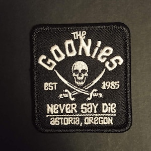 Peut inclure: Écusson carré noir avec texte et graphiques brodés en blanc. Le texte indique "THE GOONIES" au-dessus d'une tête de mort et d'épées croisées, avec "EST 1985" et "NEVER SAY DIE ASTORIA, OREGON" en dessous. L'écusson a une bordure cousue.