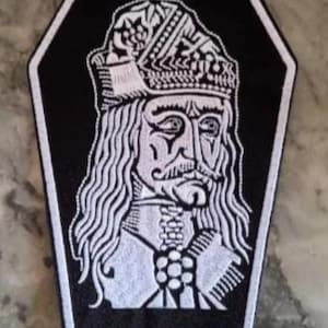 Peut inclure: Écusson brodé noir et blanc en forme de cercueil. Le patch présente un portrait détaillé d'un homme aux longs cheveux, d'une moustache et d'un chapeau décoratif. Le portrait est blanc sur fond noir.