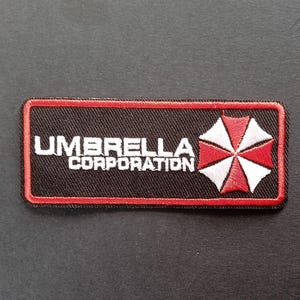 Może przedstawiać: Prostokątna haftowana naszywka z czarnym tłem i czerwoną obwódką. Napis UMBRELLA CORPORATION jest wyhaftowany białymi wielkimi literami po lewej stronie, a po prawej znajduje się czerwono-białe logo w kształcie parasola.