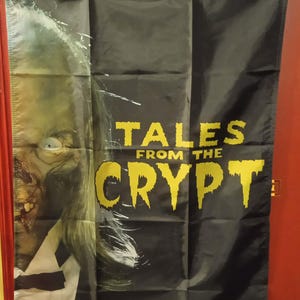 Peut inclure: Bannière noire avec le Gardien de la Crypte de Tales from the Crypt. La bannière a un fond noir avec le visage du Gardien de la Crypte à gauche et le logo de l'émission en jaune à droite. La bannière a des œillets.
