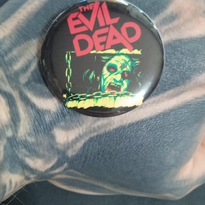 Puede incluir: Un botón redondo y negro con las palabras "THE EVIL DEAD" en letras rojas y blancas. El botón presenta un gráfico verde y amarillo de una cara de zombi. Un artículo de colección.