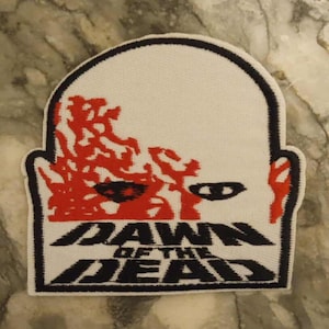 Może przedstawiać: Biała naszywka z czarną obwódką z czarnym napisem "DAWN OF THE DEAD". Naszywka przedstawia grafikę twarzy zombie z czerwonymi detalami.