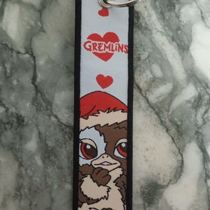 May include: A rectangular keychain featuring a Gremlins character wearing a Santa hat. The keychain has a light gray background with red hearts and the word "Gremlins" in a heart. The edges are black, and it has a silver key ring.