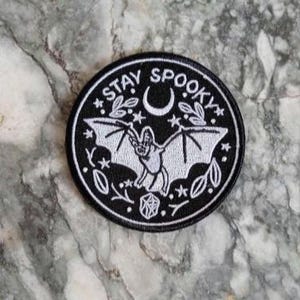 Peut inclure: Écusson rond noir et blanc avec l'inscription "STAY SPOOKY". Il présente une chauve-souris, un croissant de lune, des étoiles et des éléments végétaux. Idéal pour les vêtements ou accessoires.