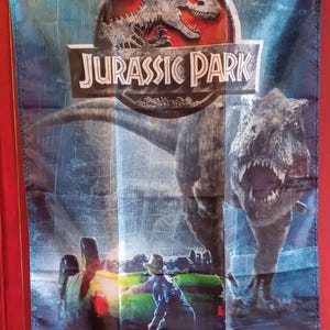 Puede incluir: Una pancarta rectangular de tela con el logotipo de Jurassic Park en la parte superior, con un gran Tiranosaurio Rex sobre una persona cerca de un vehículo. El diseño utiliza tonos azul oscuro y negro con ojales metálicos en las esquinas.