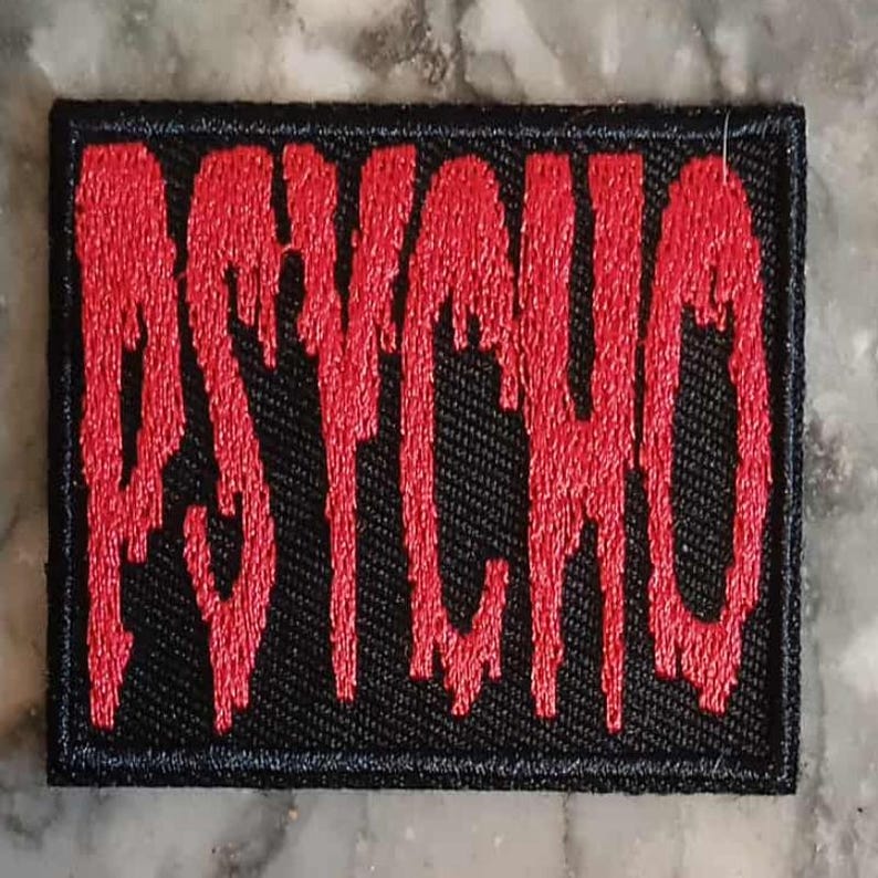 May include: A rectangular black patch with the word "PSYCHO" embroidered in dripping red letters. The patch has a black border and is likely designed to be sewn or ironed onto clothing or accessories.