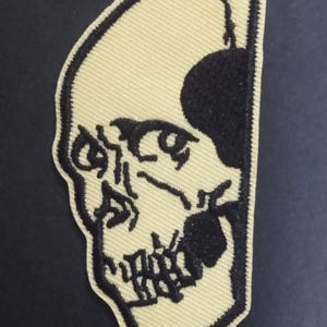 Könnte beinhalten: Gestickter Aufnäher mit einem halben Schädel-Design. Der Schädel ist in schwarzem Faden auf cremefarbenem Hintergrund dargestellt. Der Aufnäher hat einen schwarzen Rand und ist für Kleidung oder Accessoires geeignet.