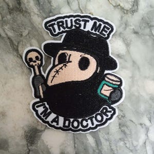 Peut inclure: Écusson brodé représentant un médecin de la peste de dessin animé avec un chapeau et un masque noirs, tenant un bâton surmonté d'un crâne et un objet vert et blanc. Le patch porte le texte "TRUST ME" au-dessus et "I'M A DOCTOR" en dessous, le tout en noir.