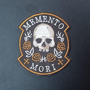 Pode incluir: Um emblema bordado circular com fundo preto apresentando um crânio branco no centro, rodeado por quatro rosas cor de laranja e detalhes de folhas brancas. O texto MEMENTO MORI está bordado a branco, com duas pequenas cruzes cor de laranja.