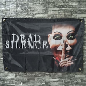 May include: Black flag featuring the text "DEAD SILENCE" in white block letters. The flag also displays a close-up of a doll's face with green eyes, a finger over its lips, and red lipstick.