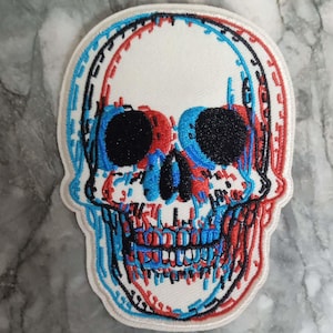 Peut inclure: Écusson brodé en forme de crâne avec un effet 3D. Le crâne présente des coutures rouges, bleues et noires sur un fond blanc. Le patch a un design unique et superposé, idéal pour les vêtements ou accessoires.