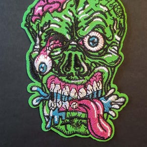 Peut inclure: Écusson brodé représentant une tête de zombie verte avec un cerveau rose, des yeux bleus et une langue rose. Le motif comprend des détails blancs, noirs et bleus, avec une bordure verte. Pour vêtements ou accessoires.