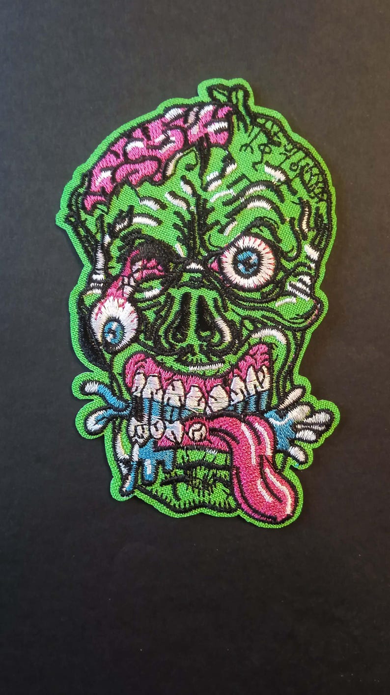 Pode incluir: Patch bordado com uma cabe&ccedil;a de zumbi verde com c&eacute;rebro rosa, olhos azuis e l&iacute;ngua rosa. O design inclui detalhes brancos, pretos e azuis, com uma borda verde. Para roupas ou acess&oacute;rios.