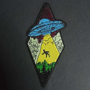 Peut inclure: Écusson brodé en forme de losange avec un OVNI bleu émettant un rayon de lumière jaune, enlevant une silhouette. Bordure noire, forêt en bas et ciel bordeaux.