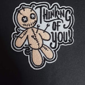 Può includere: Toppa ricamata con un bambolotto voodoo beige con cuciture e spilli neri. Il testo "Thinking of You!" è in nero, con uno stile stilizzato e un contorno bianco. Bordo nero.