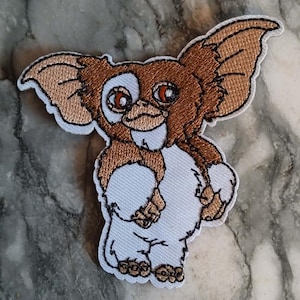 May include: Embroidered patch depicting a cartoon Gremlin character. The patch has a white background with a brown and tan Gremlin design. The Gremlin has large ears, red eyes, and a mischievous grin. The patch is outlined in black.