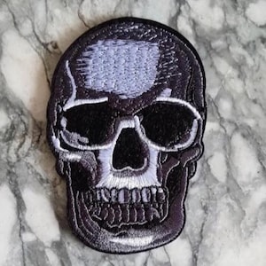 Può includere: Toppa ricamata a forma di teschio in bianco e nero. Il teschio presenta cuciture dettagliate, tra cui orbite, cavità nasale e denti. La toppa ha un bordo nero e una parte superiore grigio chiaro. Ideale per abbigliamento o accessori.