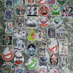 Op de afbeelding: Een verzameling kleurrijke Ghostbusters-stickers met personages, logo's en citaten uit de film. Ontwerpen omvatten de Stay Puft Marshmallow Man, Slimer en het Ghostbusters-logo.